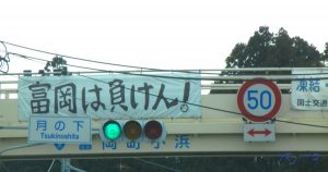 2016年3月11日に撮影した、福島県富岡町の写真です。撮影場所は、国道6号線の、月の下交差点。歩道橋に吊るされた横断幕に「富岡は負けん！」の太い字が躍ります。これからのライフデザインを提案するブログ「みらいメモ：暮らしと仕事を200字で」の記事「3.11　福島、Fukushima、忘れない」のOGP画像です。企画制作・矢嶋剛（綴り手　矢嶋ストーリーtokyo）。